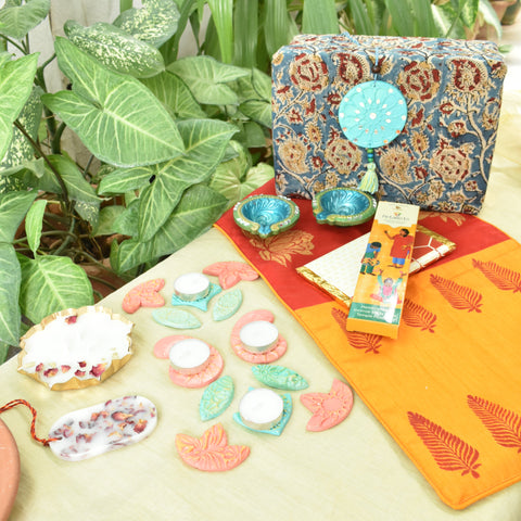 Decorative items including a kalamkari fabric box, agarbatti box, wax tablets and candles and decorative rangoli on a surface with plants in the background.